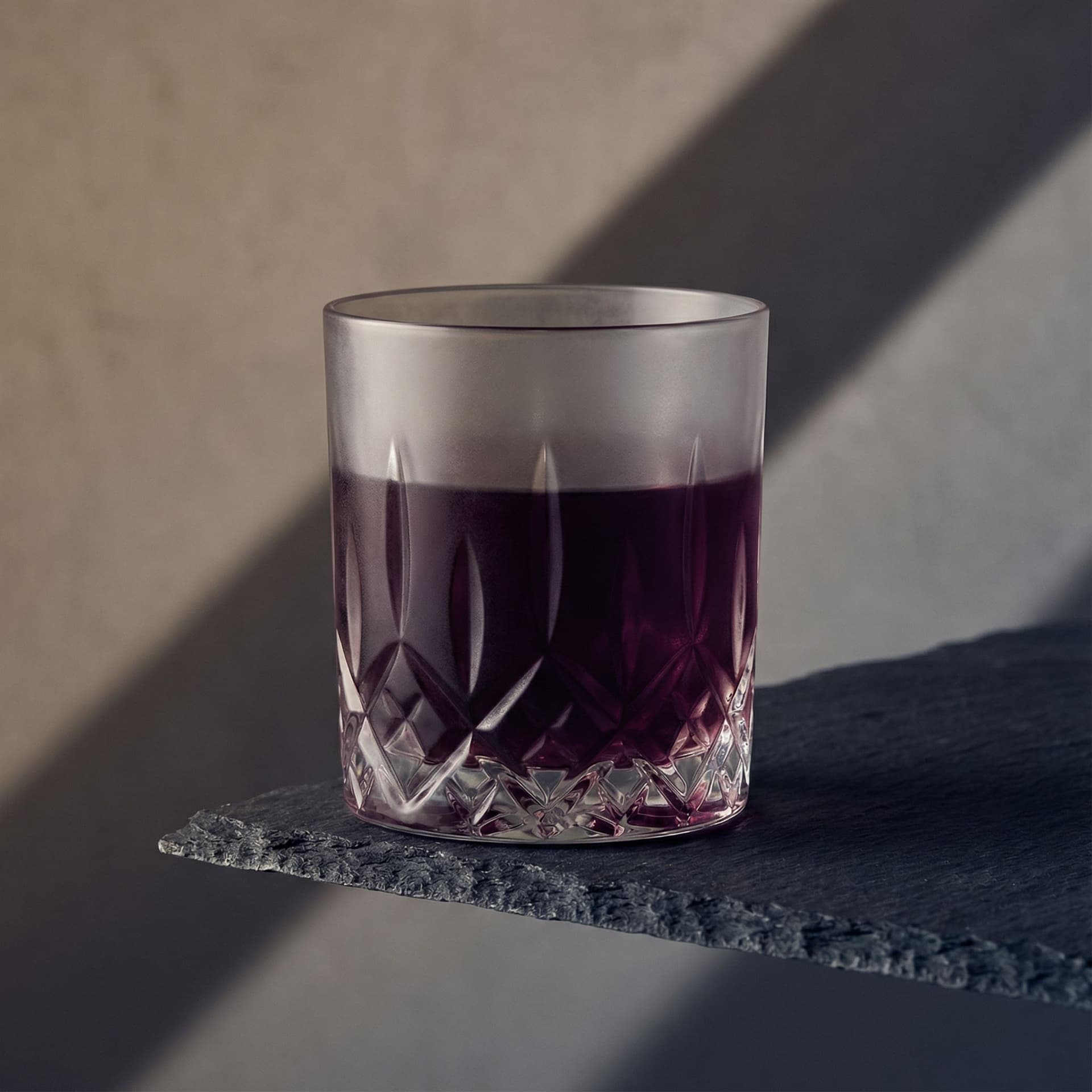 a heavy, low-cut crystal tumbler with a dark plum-toned negroni, placed perfectly aligned with the edge of a rough slate surface, its symmetry disrupted by a single sharp diagonal shadow cast from a narrow beam angled from the side