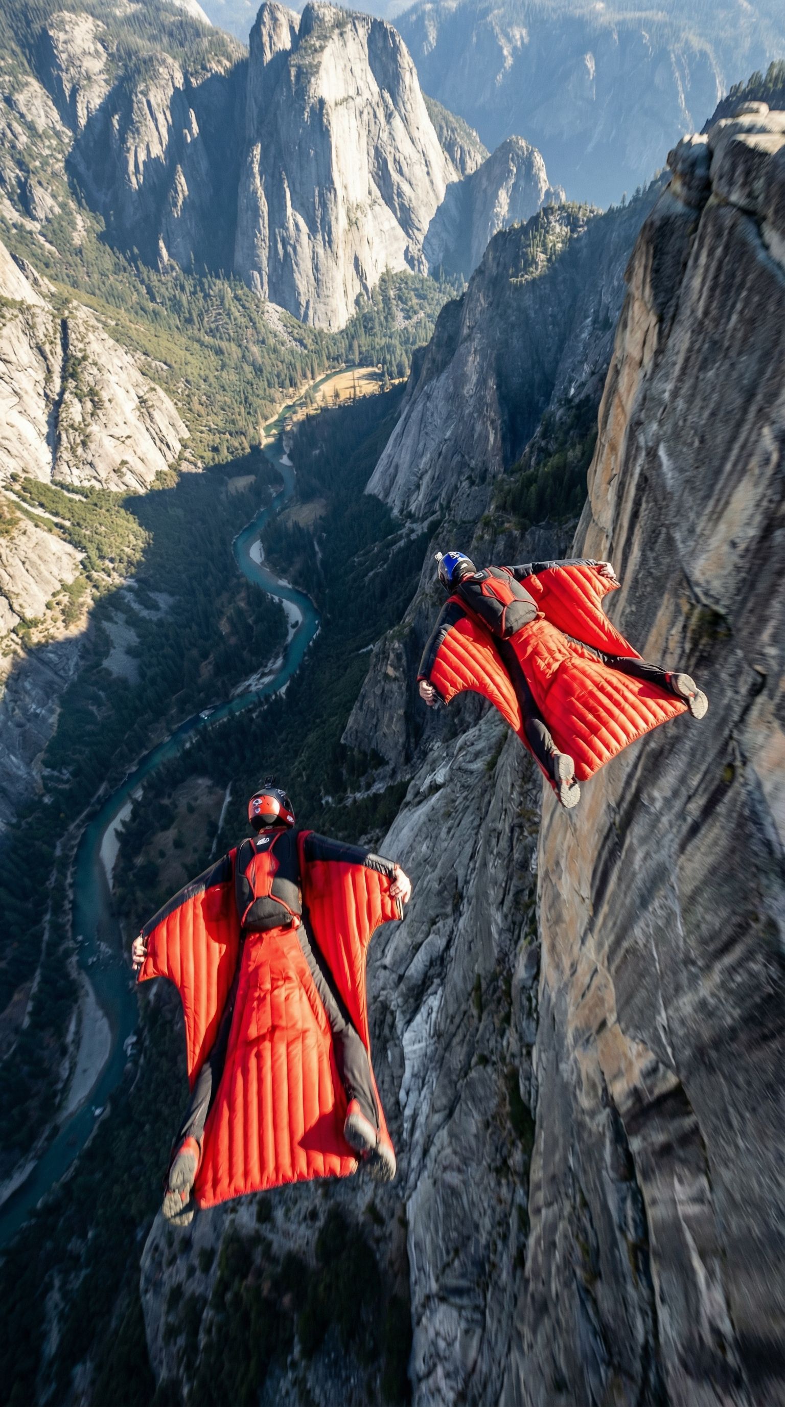 A high-detail realistic shot of two wingsuit flyers soaring down a massive mountain valley, wearing bright red wingsuits, dramatic aerial perspective, huge cliffs and a winding blue river below, crisp lighting, intense sense of speed and freedom.
